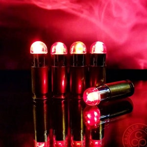 Puede incluir: Cinco bombillas LED rojas en fila, cada una encerrada en una bombilla de vidrio transparente y una carcasa de latón. Las bombillas están encendidas y emiten un brillo cálido. Las bombillas se reflejan en una superficie brillante.