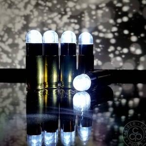 Puede incluir: Cinco luces LED blancas con forma de bala con una carcasa negra y una base dorada. Las luces están dispuestas en una fila sobre una superficie reflectante. Las luces están encendidas e iluminan la superficie.