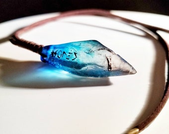 May include: A blue glass pendant with a pointed tip, hanging from a brown leather cord. The pendant has a dark inscription that reads "JEVON".