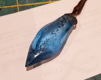 May include: A blue glass pendant with a brown cord. The pendant has a pointed tip and is inscribed with the text "The Sword".