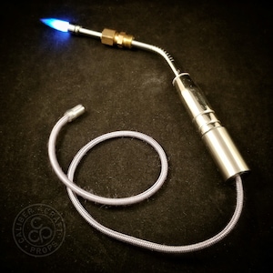 May include: A silver torch with a blue flame and a flexible hose. The torch is attached to a silver cylinder with a brass fitting. The hose is coiled on a black surface.