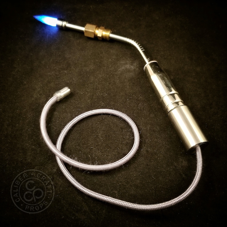 May include: A silver torch with a blue flame and a flexible hose. The torch is attached to a silver cylinder with a brass fitting. The hose is coiled on a black surface.