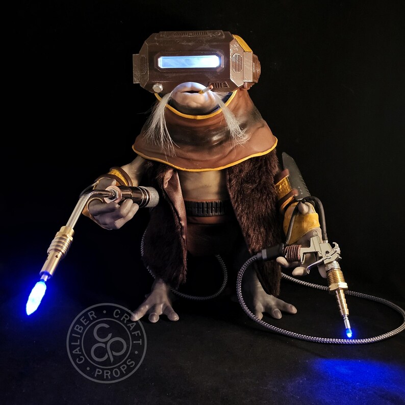 May include: A detailed prop figure of a creature with a brown and tan outfit, a fur-lined coat, and a welding mask with a blue light. The figure holds two welding tools with glowing blue tips. The logo "CALIBER CRAFT PROPS" is visible.