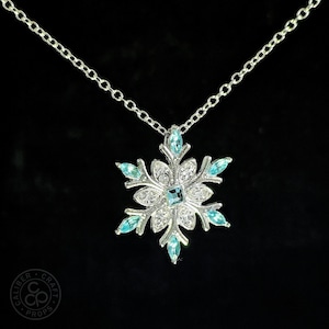 May include: Silver-toned snowflake pendant necklace with a delicate chain. The snowflake design features clear and light blue crystals, with a square blue crystal at the center. The necklace is set against a black background.