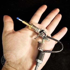 May include: A miniature silver and gold prop gun with a black cable and a braided gray cord. The prop gun has a blue tip and a brown wrapped handle. The logo "Caliber Craft Props" is visible in the lower left corner.