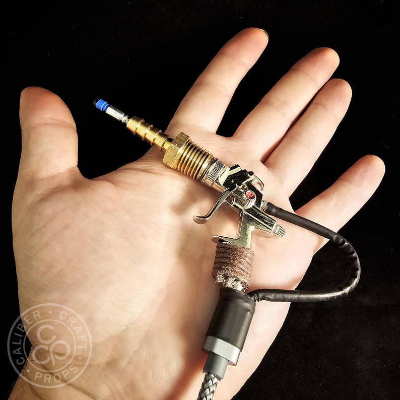 May include: A miniature silver and gold prop gun with a black cable and a braided gray cord. The prop gun has a blue tip and a brown wrapped handle. The logo "Caliber Craft Props" is visible in the lower left corner.