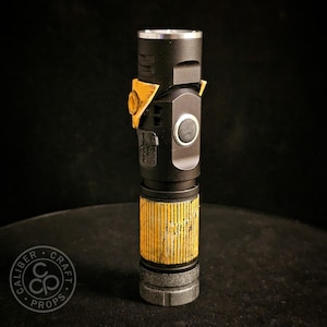 May include: A black cylindrical torch prop featuring a weathered yellow grip section with vertical grooves, a metallic silver top rim, a circular side button, and a yellow triangular accent piece near the top with a small circular cutout.