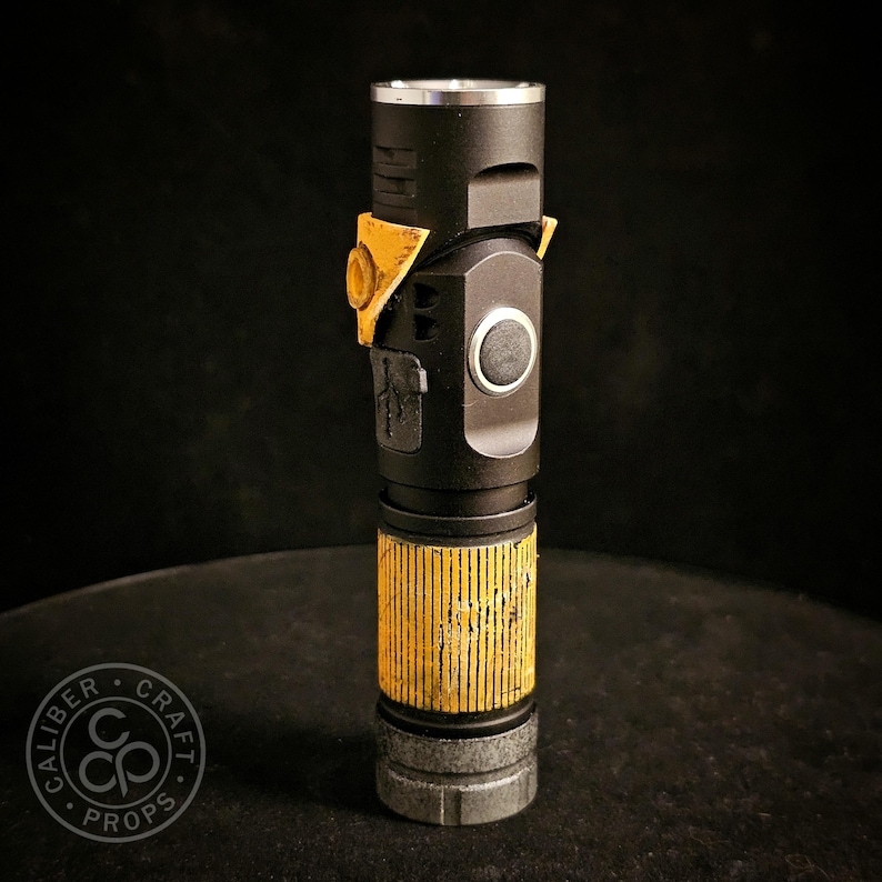 May include: A black cylindrical torch prop featuring a weathered yellow grip section with vertical grooves, a metallic silver top rim, a circular side button, and a yellow triangular accent piece near the top with a small circular cutout.