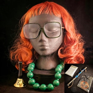 May include: A mannequin head with a red wig and large square eyeglasses. A green beaded necklace is around the neck. A small gold bell, a notebook, a wand, and a pair of copper-colored earrings are also visible.