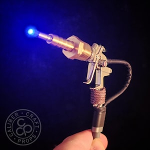 May include: A steampunk-style prop gun with a blue LED light at the barrel. The gun features brass and silver-colored metal components, a trigger, and a wrapped handle. A black cable extends from the base.