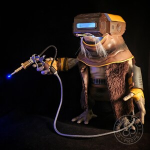 May include: A detailed prop of a science fiction character with a brown and yellow helmet, fur-lined coat, and a futuristic ray gun. The ray gun has a blue light emanating from the barrel. The character has a long white beard and is holding the ray gun.