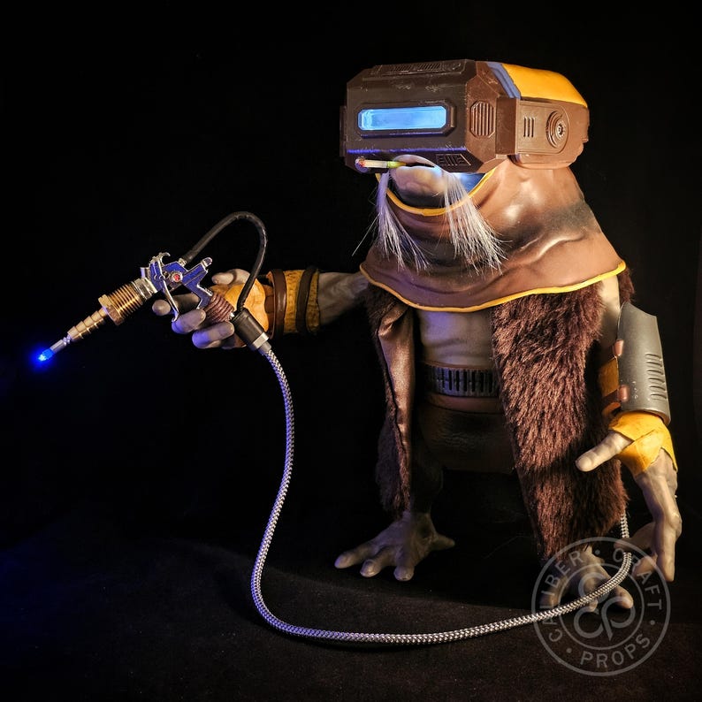 May include: A detailed prop of a science fiction character with a brown and yellow helmet, fur-lined coat, and a futuristic ray gun. The ray gun has a blue light emanating from the barrel. The character has a long white beard and is holding the ray gun.