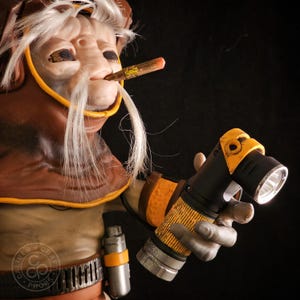 May include: A detailed figurine of a creature with white hair and a cigar in its mouth, wearing a brown and yellow hood. The figure holds a black and yellow flashlight in its hand. A small silver canister is attached to the belt of the figure.