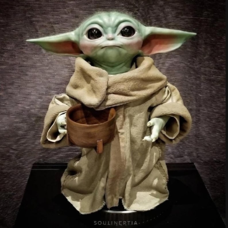Puede incluir: Figura detallada de un personaje con grandes ojos negros, piel verde y orejas grandes, que lleva una t&uacute;nica marr&oacute;n y sostiene un peque&ntilde;o cuenco de madera. La figura est&aacute; sobre una base negra.