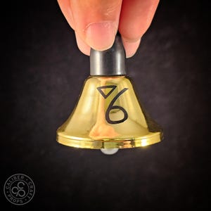 May include: A small, gold-colored bell with a black top and a black number 6 symbol. The bell is held by a hand against a dark background. The bell is approximately 2 inches tall.