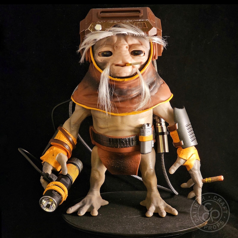 May include: A detailed figurine of a creature with light skin, white hair, and a brown hood. It wears a brown helmet, orange wrist guards, and a dark belt with a silver canister. One hand holds a yellow and black flashlight, while the other holds a small cigar.