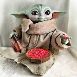May include: A detailed figurine of a character from a popular science fiction series. The figure is light green with large black eyes, wearing a tan robe. It holds a box with Japanese text and a wooden bowl filled with red candies.
