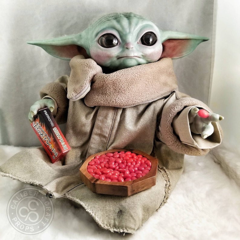 May include: A detailed figurine of a character from a popular science fiction series. The figure is light green with large black eyes, wearing a tan robe. It holds a box with Japanese text and a wooden bowl filled with red candies.