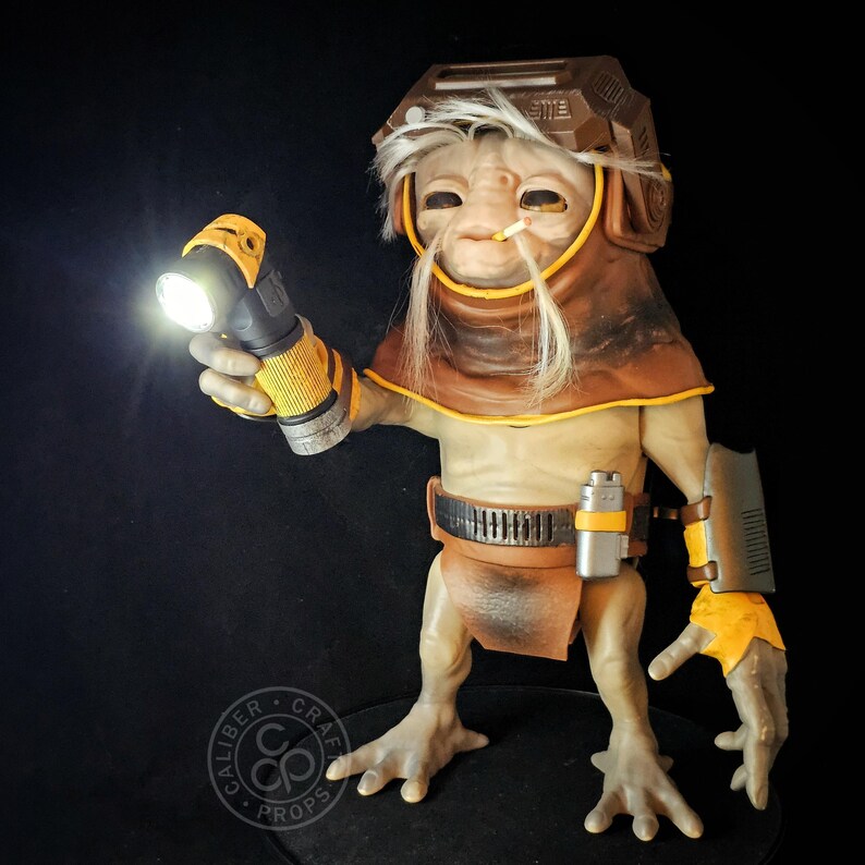 May include: A detailed creature figurine with light tan skin, wearing a brown hood and helmet, holding a bright, illuminated yellow and black flashlight. The figure features long white hair, a small cigarette in its mouth, and a utility belt.