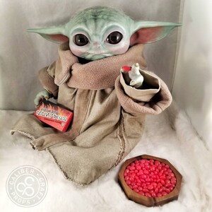 May include: A green-skinned doll with large eyes, wearing a tan robe and holding a small box of cinnamon candy. A wooden bowl filled with red jelly beans sits nearby. The doll has large ears and is a character from a popular science fiction franchise.