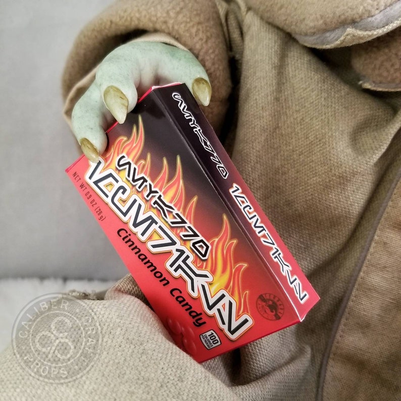 May include: A red and black box of "WYK770 Cinnamon Candy" is held in a green hand with long nails. The box features flames and text in a stylized font. The candy box is 0.9 oz (26g).