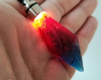 Op de afbeelding: Een ketting met een rode en blauwe kristallen hanger met een lichtje erin. De hanger heeft de vorm van een diamant en heeft een zilveren metalen bovenkant.