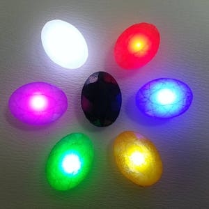 May include: Seven oval-shaped stones with different colors and textures, each emitting a different colored light. The stones are arranged in a circle, with a black stone in the center.