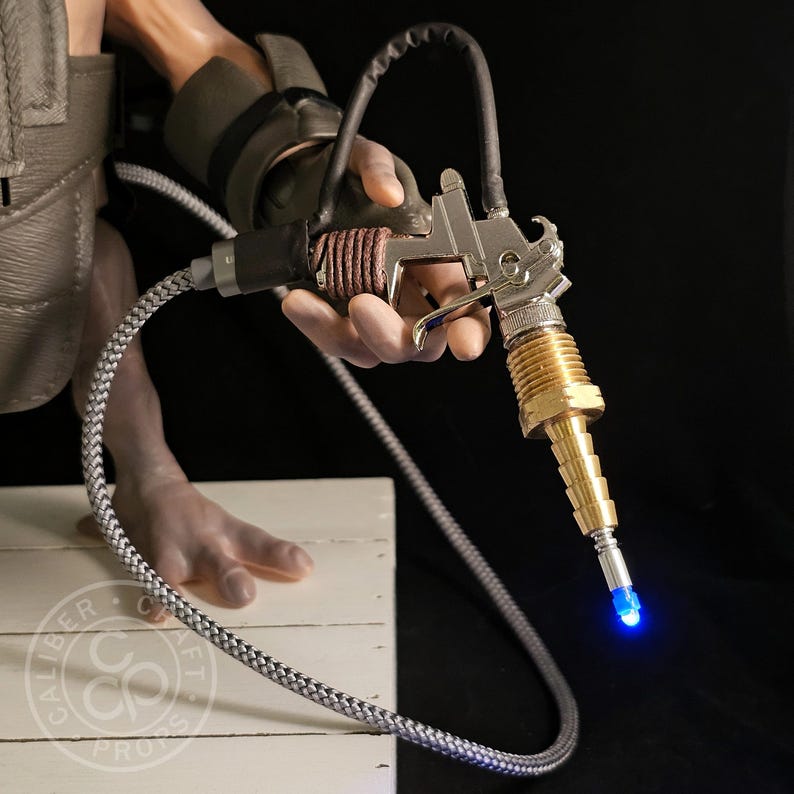 May include: A prop gun with a silver body, brass nozzle, and a blue light at the tip. The gun is held by a person wearing a dark sleeve and a gray vest. A braided gray and white hose is attached to the gun.