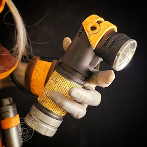 May include: A handheld torch with a black body and a yellow, textured grip section. The head of the torch is angled, featuring a yellow frame and a metallic silver rim around the lens. The design includes weathered, distressed details.