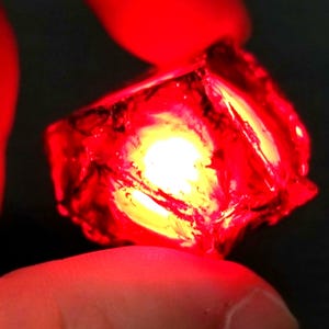 May include: A red, faceted, cube-shaped crystal with a bright light shining through it.