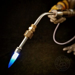 May include: A close-up of a silver and brass torch with a blue flame. The torch is held by a hand wearing a glove. The torch is labeled "Caliber Craft Props".