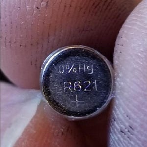 以下が含まれることがあります： 小さな銀色のボタン電池のクローズアップ。電池の表面には「0% Hg」と「LR621」の文字が印刷されています。プラス記号も表示されています。電池は2本の指で挟まれています。