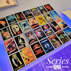 May include: A collection of rectangular holographic stickers featuring iconic horror movie posters including The Texas Chainsaw Massacre, Friday the 13th, Child's Play, Poltergeist, The Rocky Horror Picture Show, They Live, and Scanners.