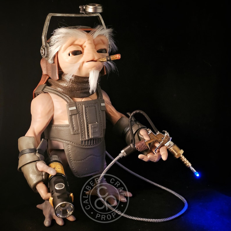 May include: A detailed bust of an alien character with a white beard, wearing a headset and a utility vest. The character holds a lit cigar and a futuristic ray gun with a blue light. A flashlight is also visible. The bust is a prop.