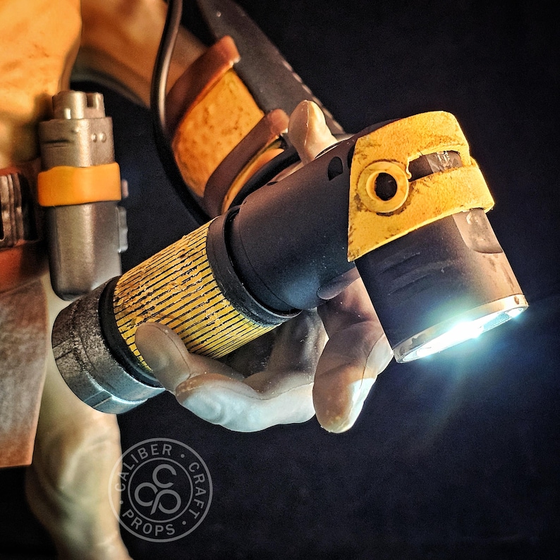 May include: A black and yellow torch with a textured grip and a bright LED beam. The torch features a weathered finish with yellow accents on the head and body. A small cylindrical canister with a yellow band is attached to the side.