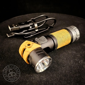 May include: A black and yellow torch with a textured grip and a silver metallic bezel, accompanied by a black USB charging cable. The torch features a side-mounted light head and a weathered finish on the yellow components.