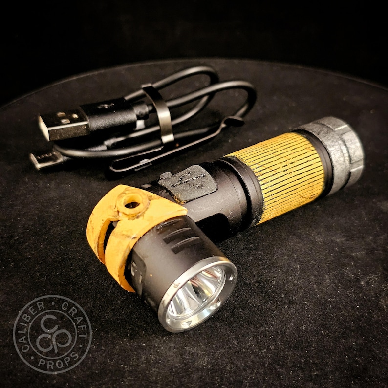 May include: A black and yellow torch with a textured grip and a silver metallic bezel, accompanied by a black USB charging cable. The torch features a side-mounted light head and a weathered finish on the yellow components.