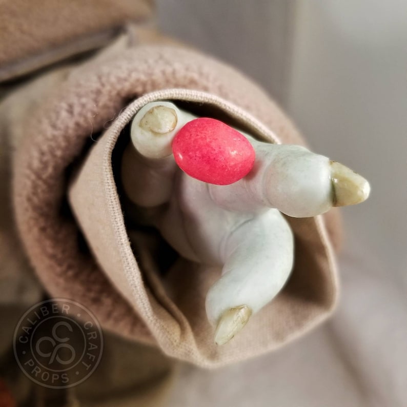 May include: A close-up of a pale green hand with long, off-white fingernails, holding a bright red, oval-shaped object. The hand is emerging from a tan fabric sleeve. The image has a soft focus.