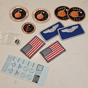 Op de afbeelding: Een verzameling patches, stickers en een speld. Patches zijn onder meer de Amerikaanse vlag, het woord "ARES" met een planeetontwerp en namen als "LEWIS" en "WATNEY". Stickers bevatten tekst over overlevingsinstrumenten in de ruimte.