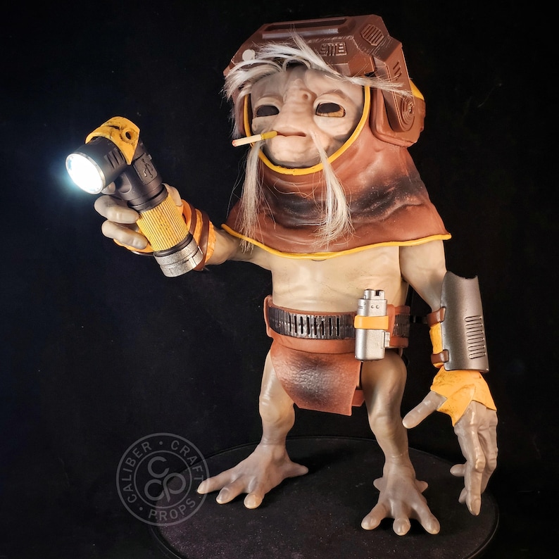 May include: A detailed figurine of a creature with wrinkled skin, wearing a brown hood and helmet, holding a lit flashlight. The figure features white hair, a small cigarette in its mouth, a dark belt with a silver canister, and a metallic arm guard.