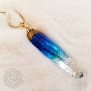 May include: A gold chain necklace with a blue and clear crystal pendant. The pendant is shaped like a long, pointed crystal. The crystal is a gradient of blue, fading to clear at the tip.