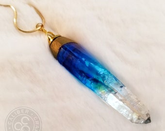 May include: A gold chain necklace with a blue and clear crystal pendant. The pendant is shaped like a long, pointed crystal. The crystal is a gradient of blue, fading to clear at the tip.