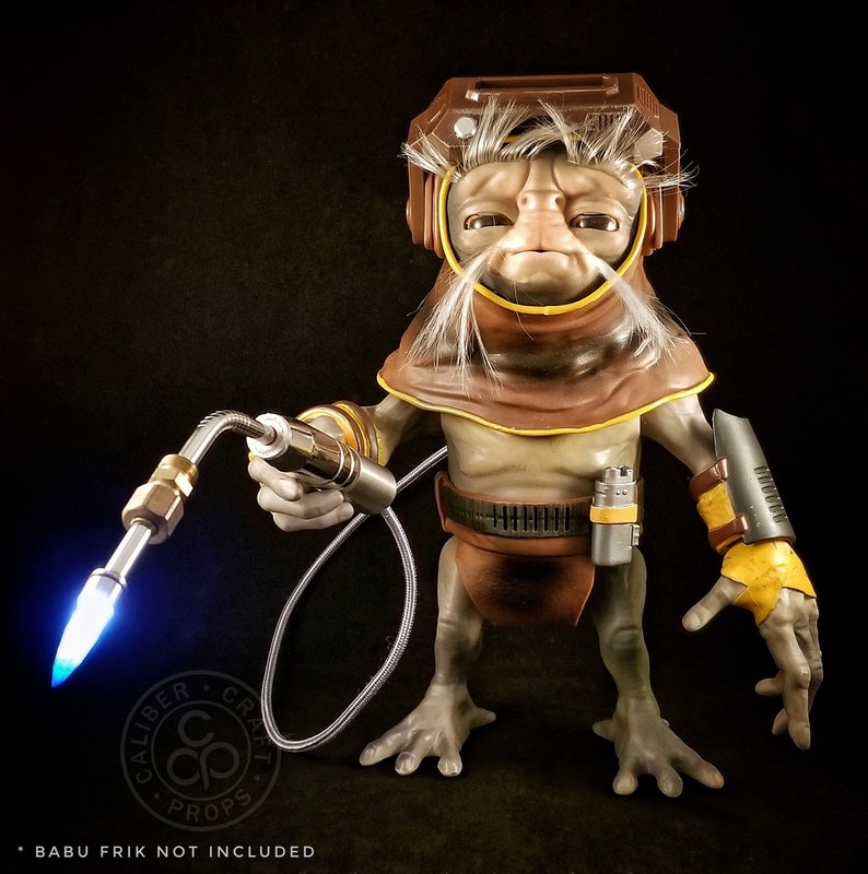 May include: A brown and gray action figure of a character from Star Wars with a welding torch. The figure is wearing a brown hooded robe and has a white beard. The figure is holding a welding torch in its right hand. The figure is standing on a black surface. "Babu Frik not included" is written at the bottom of the image.
