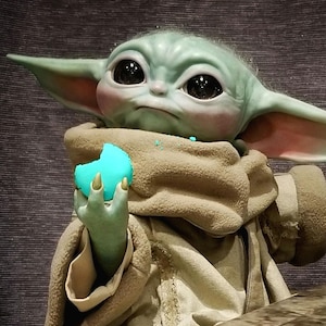 May include: A detailed figurine of a green alien-like character with large black eyes, holding a bright blue treat. The character is wearing a tan robe and has large ears. The figure is a popular character from a science fiction franchise.
