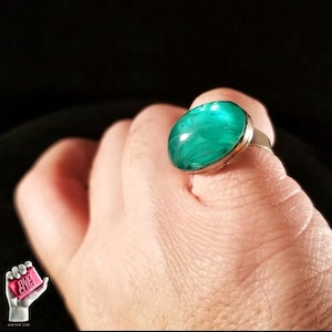 May include: A silver ring featuring a large, oval, teal gemstone. The stone has a marbled effect. The ring is set against a dark backdrop. The ring is on a finger.