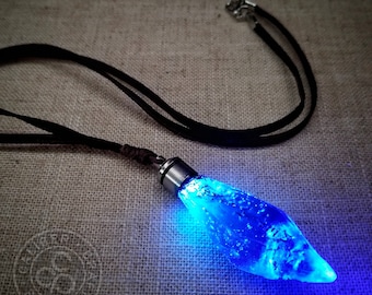May include: A blue glowing crystal pendant necklace with a black leather cord. The crystal is shaped like a teardrop and has a silver cap at the top.