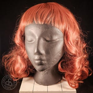 May include: A copper-colored wig with a short, fringed bang and shoulder-length curls is displayed on a gray mannequin head. The wig is styled with loose waves and sits atop a white, rectangular base. The Caliber Craft Props logo is visible.