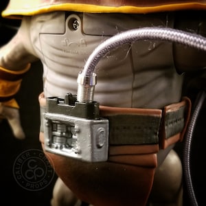 May include: A close-up of a silver and black action figure accessory. The accessory is a belt with a silver buckle and a black box with a silver detail. A purple cable is attached to the box.