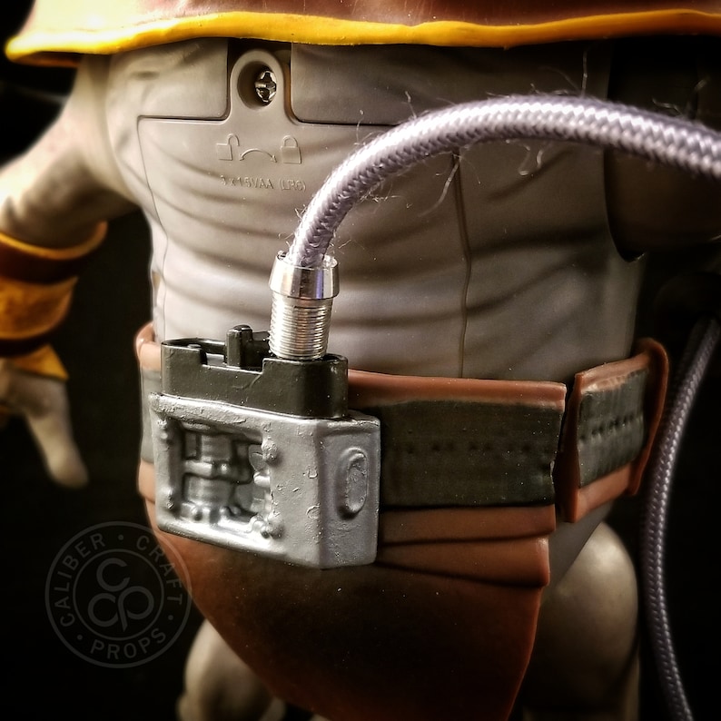 May include: A close-up of a silver and black action figure accessory. The accessory is a belt with a silver buckle and a black box with a silver detail. A purple cable is attached to the box.