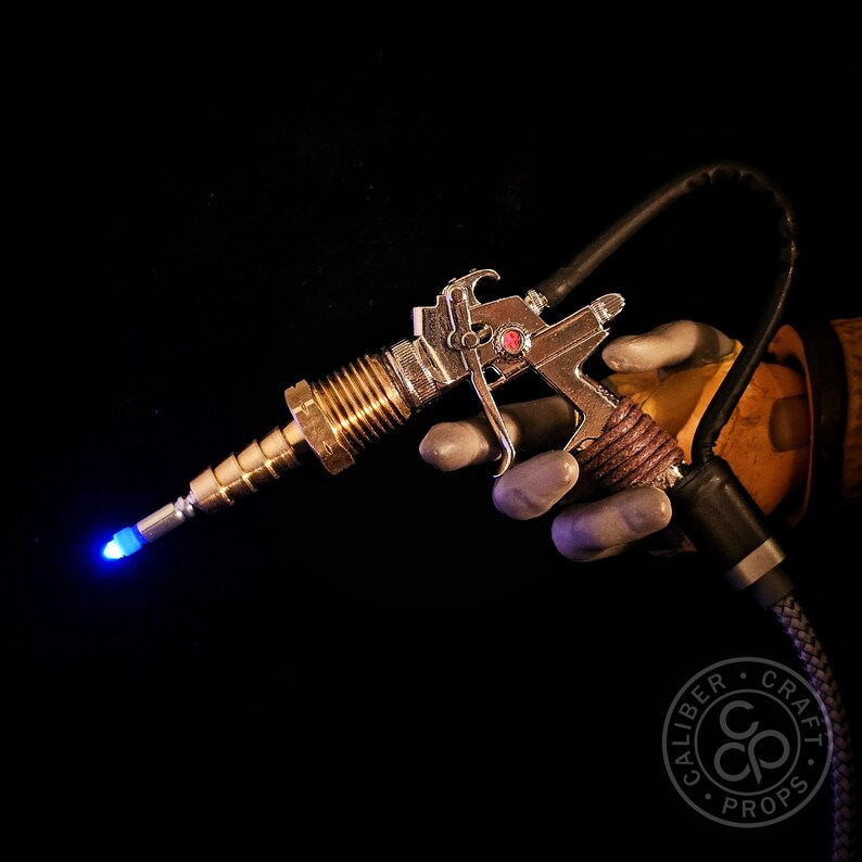 May include: A steampunk-style prop gun with a silver and brass finish, featuring a glowing blue tip. The gun is held against a black background, with a red gem detail and a coiled brown handle. A black hose extends from the gun.